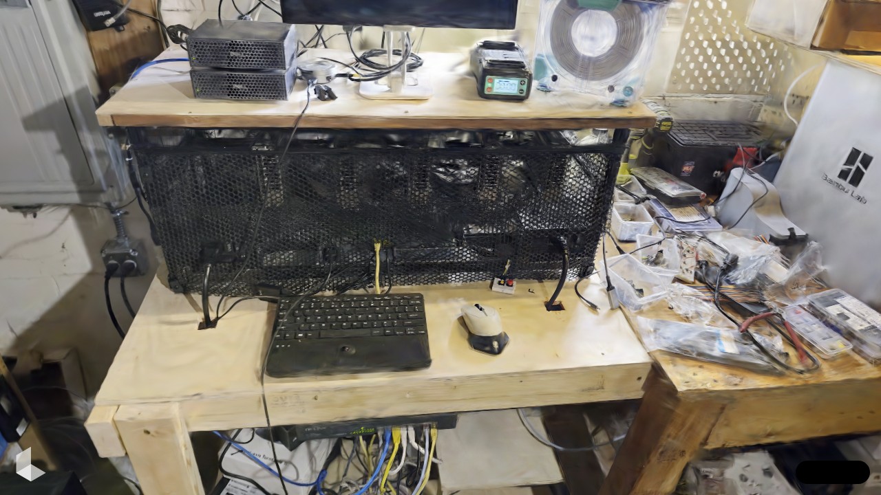 Gaussian splat capture of juicebox, an open-frame GPU rig behind black mesh on a plywood bench in the basement, keyboard and mouse in front, cabling everywhere