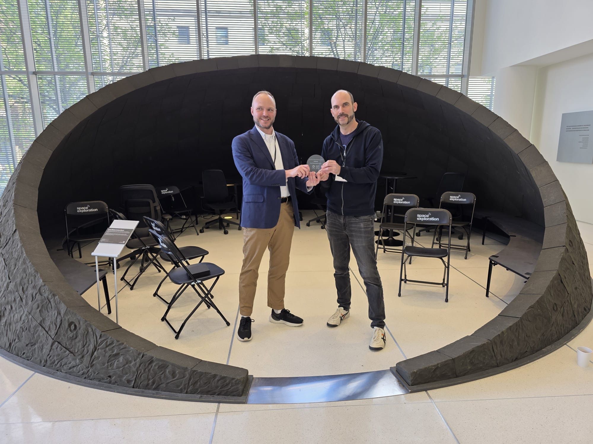 Martin and Daniel Pillis holding the award inside a dark, cave-like circular architectural installation, stackable black chairs arranged in a circle, signage nearby reading “space exploration”